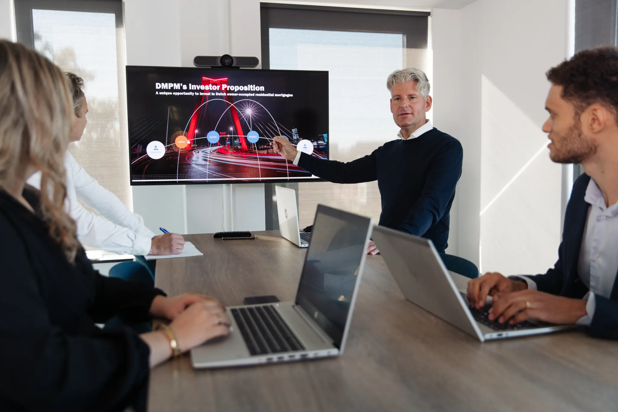 Man presenting on a tv screen. A woman and man sitting behind laptop and watching the presentation.